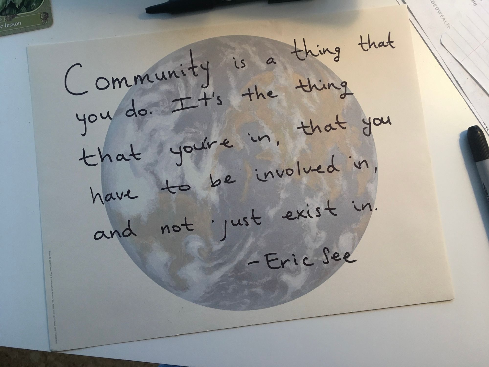 paper on white desk with some paper mess, a marker to the side and a pen above, and a corner of an oracle card. the sheet of paper has an image of the planet diffused as a background, and I wrote in sharpie: "Community is a thing that you do. IT's the thing that you're in, that you have to be involved in, and not just exist in. - Eric See"