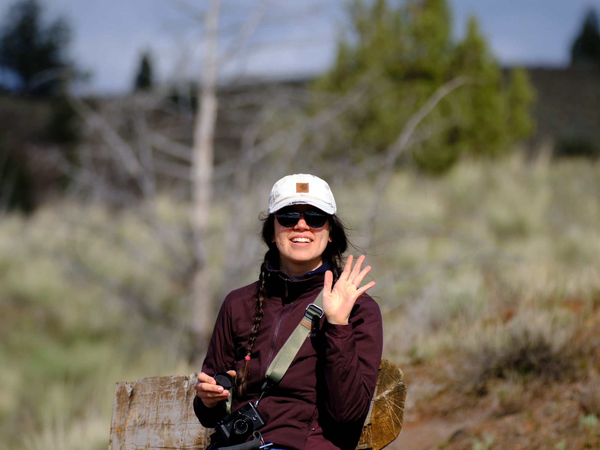 image of Cassandra wearing baseball cap with long brown-haired braid, she's wearing purple jacket and has her left hand up frozen in an open palmed wave at Nathan who's taking the photo. She has a lens cap in her other hand and a small camera strapped around her as she sits on a bench with a blurry high desert background.