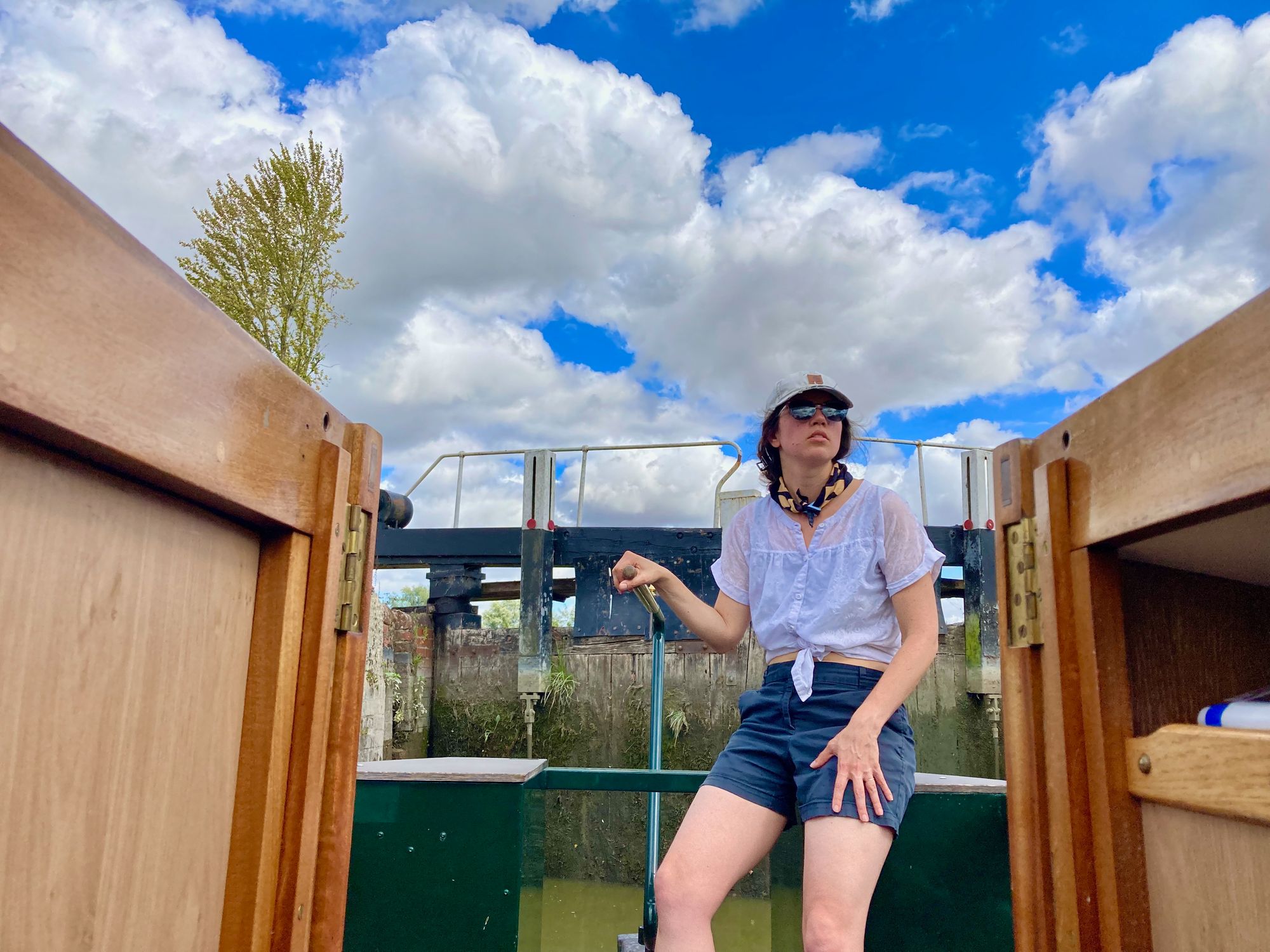 Cassandra sitting on back of canal boat with R hand on tiller, she sits looking out as she pilots, closed locks with algae and plants growing on them are right behind her, photographer has taken shot within cabin so Cassandra is framed by wooden cabin entrance and bright blue skies with puffy clouds.