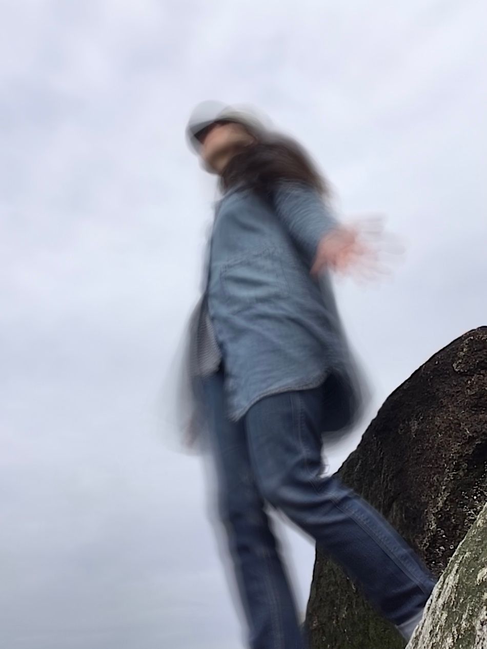 Now the camera/phone is on the ground and looking up at Cassandra as she dances. The image is a time-lapse and so Cassandra (wearing long jean button up shirt and dark jean pants) is dancing and thus blurry because of the time-lapse. She has arms to her sides with the palms facing out. She is standing in front of boulders. Behind/Above her is a blue-gray sky.