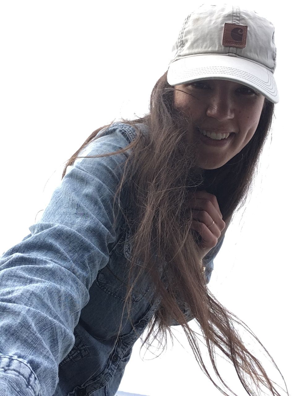 Cassandra is smiling and wearing her trusty gray Carhartt baseball cap and jean long sleeve button up sheet. Her long brown hair is waving in the wind. She is looking at camera as she holds camera. She is in the midst of placing camera/phone down on ground.