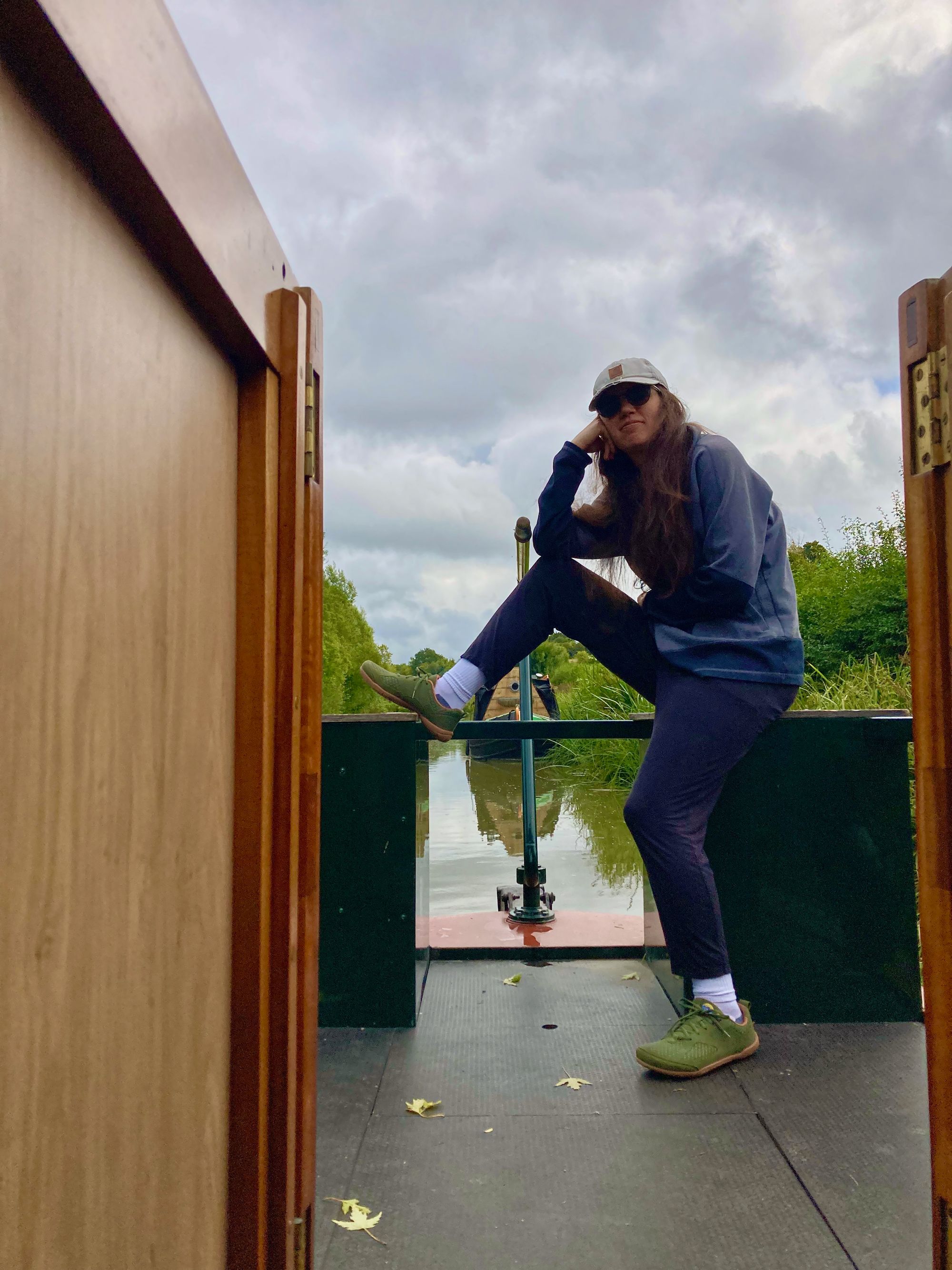 Cassandra sits with one leg on ground and one leg bent and up on back end cabinet of canal boat. She is wearing dark clothes and looks a bit glum. the boat is moored and there is greenery behind her as well as another moored canal boat. the sky is overcast.