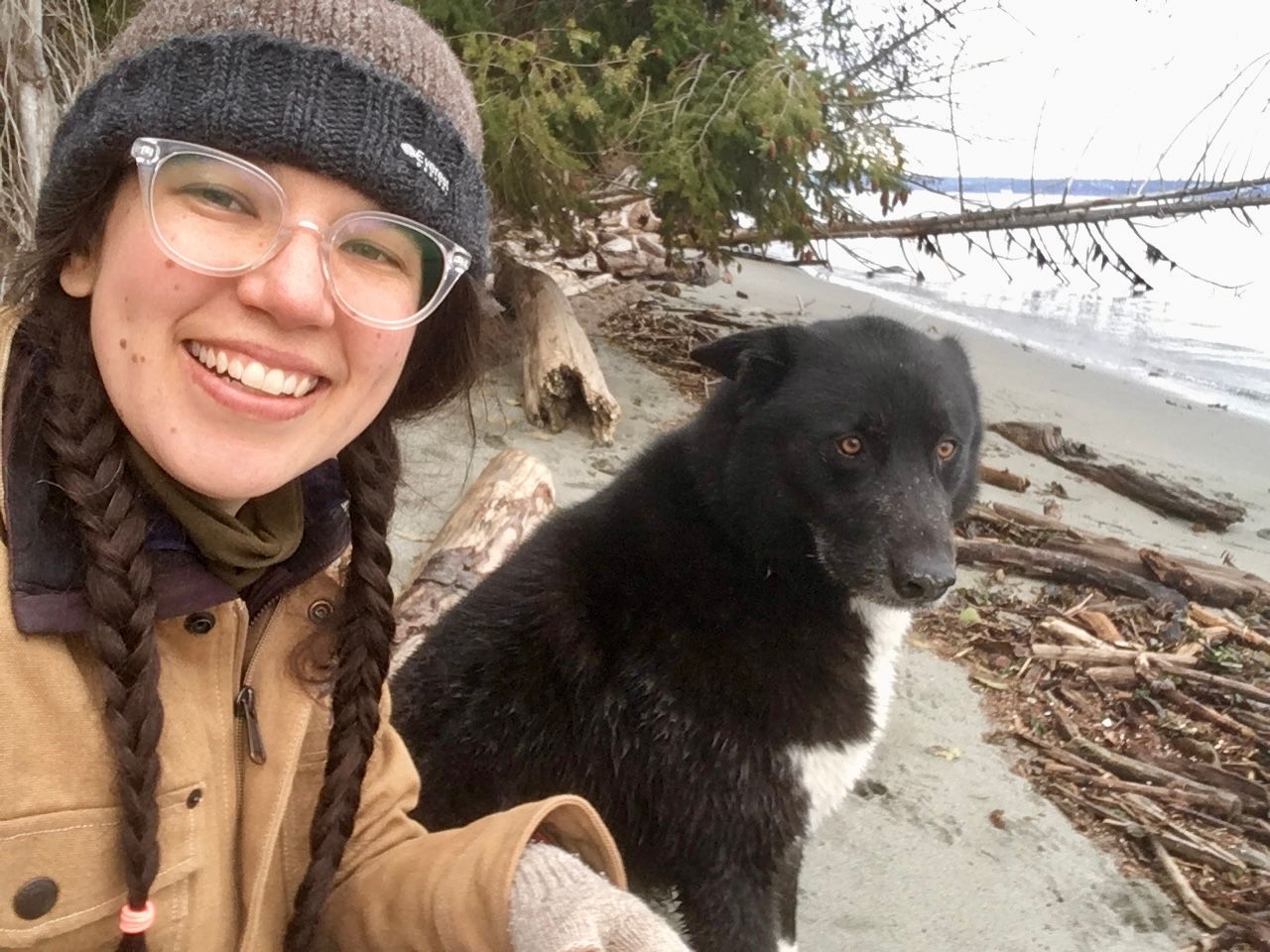 Cassandra sits smiling at camera wearing a warm hat, two braids, glasses, and jacket. Ernest is slightly behind and next to Cassandra, looking at past the camera, he is a bit shaggy from being in the water and has sand on his nose. There is lots of driftwood on the beach and trees, one fallen, into the water beyond them.