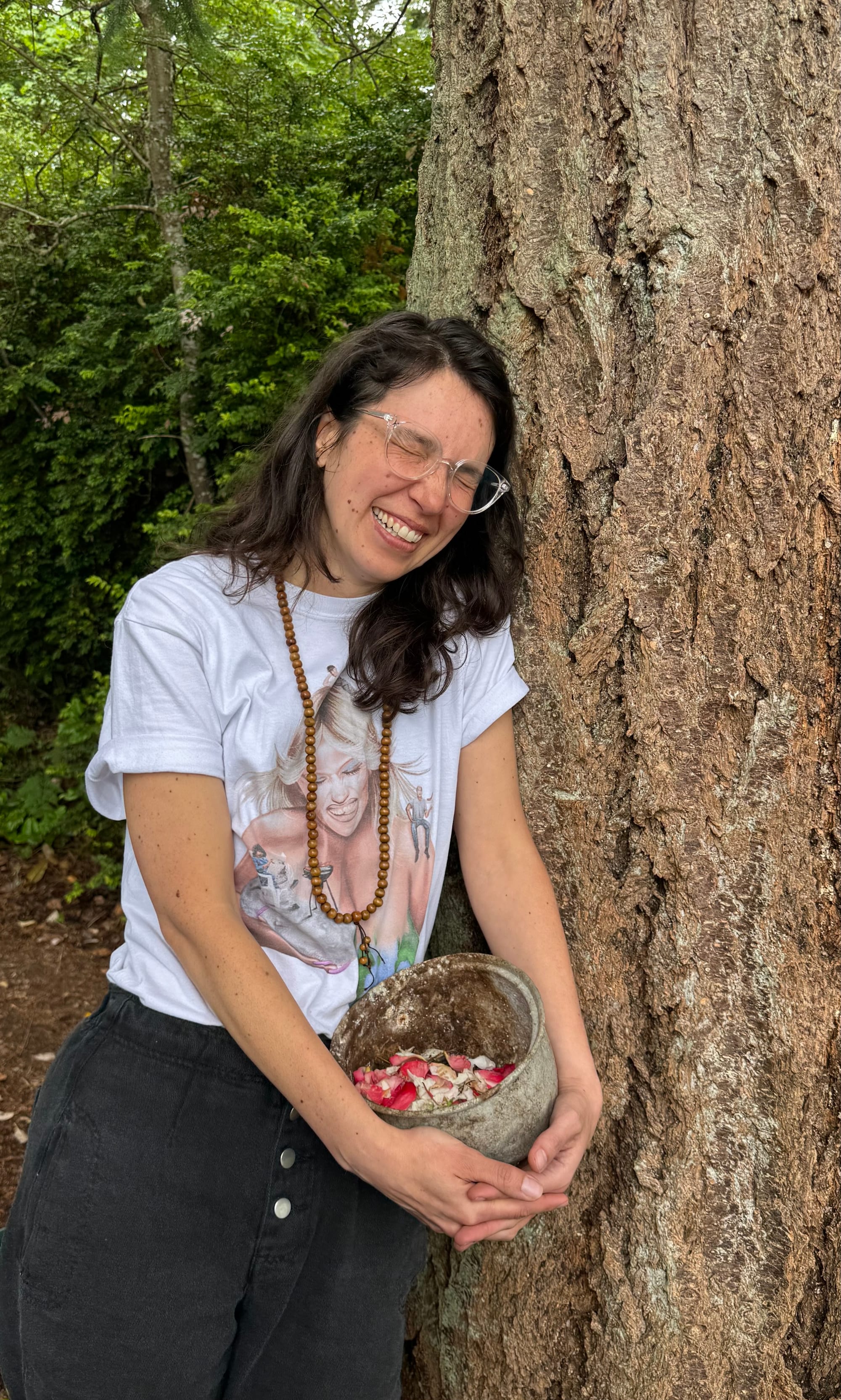 Cassandra leans against a tree while laughing, eyes closed, and holding a rusty bowl filled with white and pink rose petals, she is wearing a mala beaded necklace and a white Amyl and the Sniffers shirt with black pants
